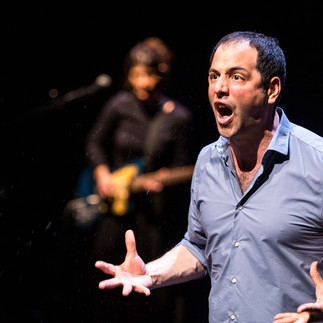 Un acteur masculin sur scène, captivant et expressif, s'adresse passionnément au public avec un musicien flou en arrière-plan, illustrant l'union de la narration et de la musique en direct.