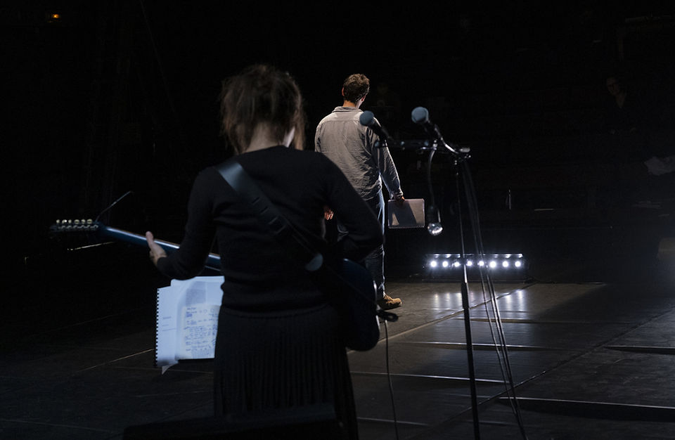 Vue de la scène vers le public, un acteur masculin de dos face à une musicienne de face, dans l'immensité de l'espace théâtral.