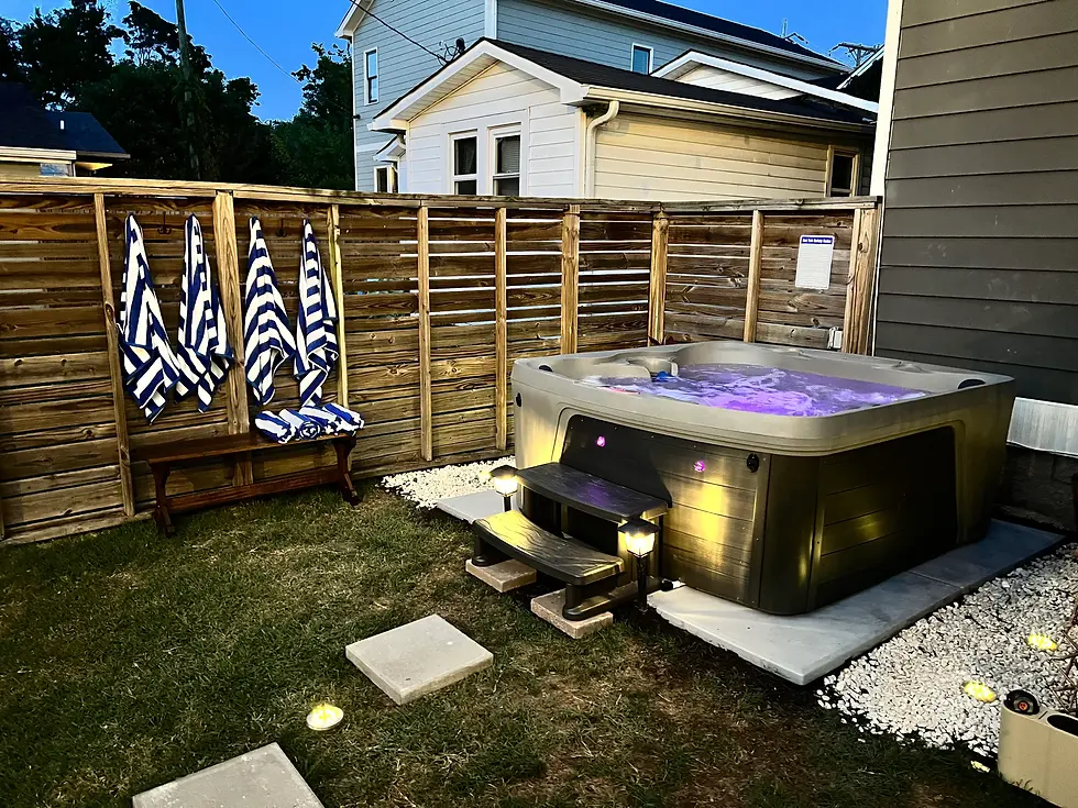 Illuminated hot tub in private backyard with wooden fence, towels, and landscape lighting near modern home