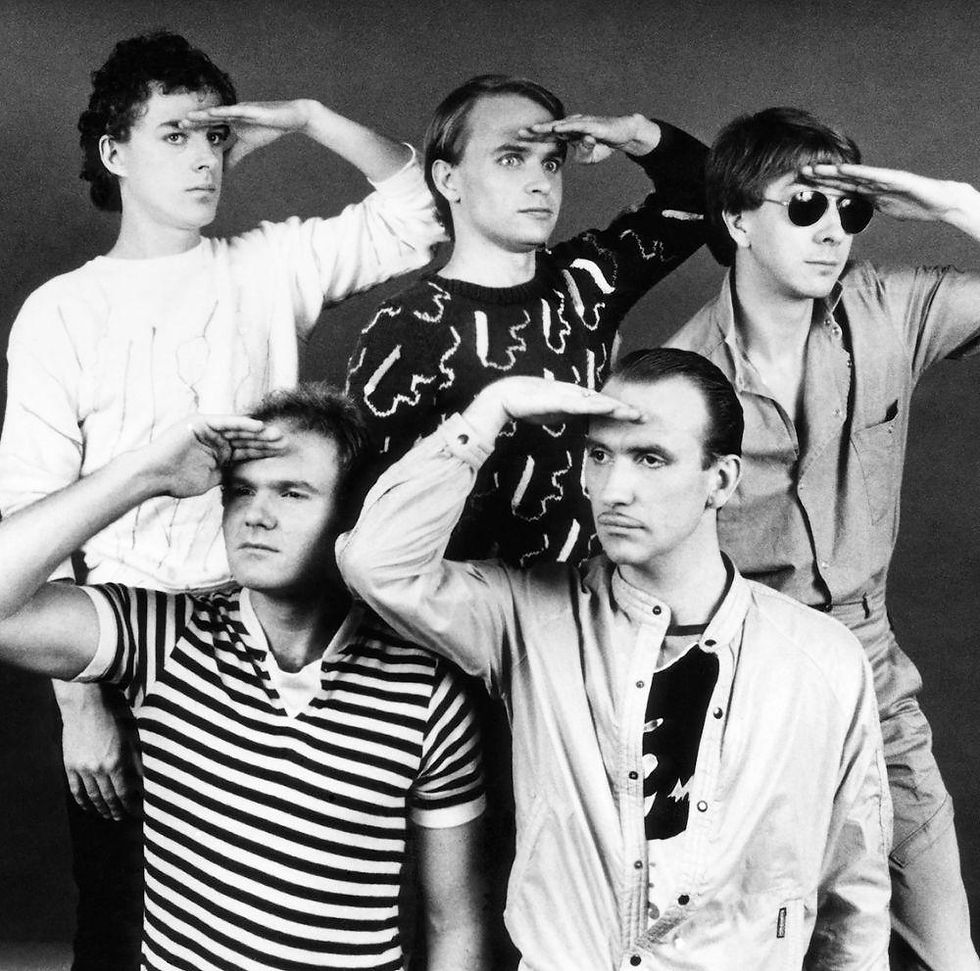 Black-and-white promotional photo of Men at Work's classic lineup from the early 1980s, with all five band members humorously saluting while looking into the distance. From left to right are Colin Hay, Greg Ham, Ron Strykert, John Rees, and Jerry Speiser.