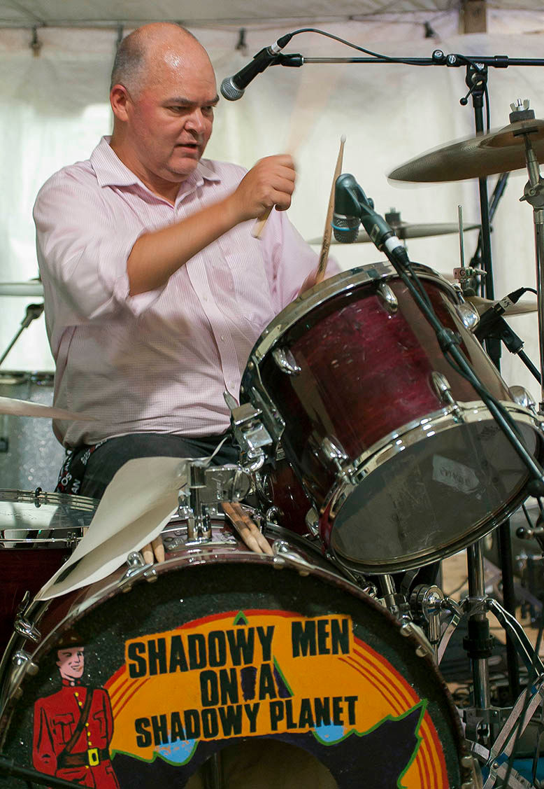 Don Pyle performs live onstage behind a drum kit featuring the logo of Shadowy Men on a Shadowy Planet. He holds drumsticks mid-performance while seated at a red drum set under stage microphones and lighting.