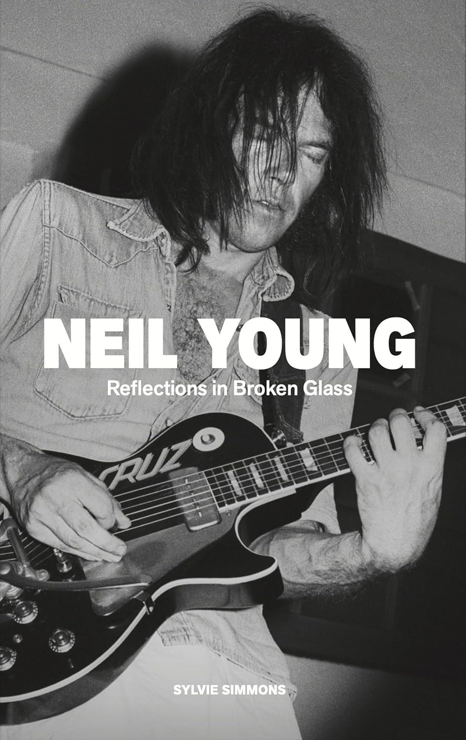 Cover of Neil Young: Reflections in Broken Glass by Sylvie Simmons, featuring a black-and-white photo of Neil Young playing a black electric guitar with long dark hair covering part of his face. The updated biography explores Neil Young's life, music, and career through new interviews and decades of rock history.