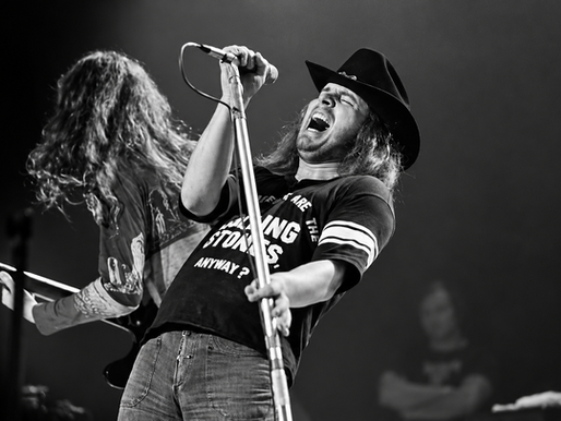 Black-and-white concert photo of a long-haired rock singer in a black cowboy hat singing passionately into a microphone onstage, with a guitarist performing behind him.