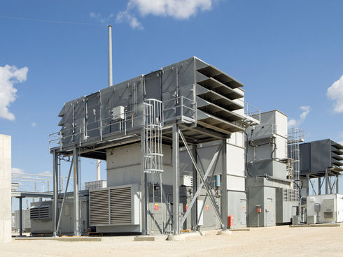 Industrial facility with large gray structures and vents, set against a blue sky with clouds. Gravel ground and some safety signs visible.