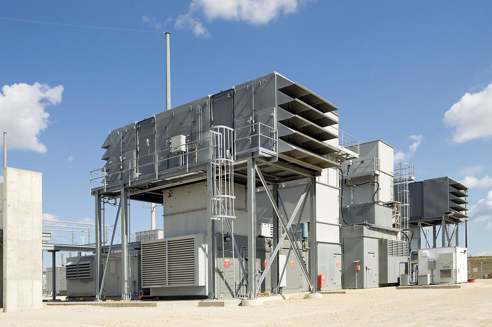 Industrial facility with large gray structures and vents, set against a blue sky with clouds. Gravel ground and some safety signs visible.