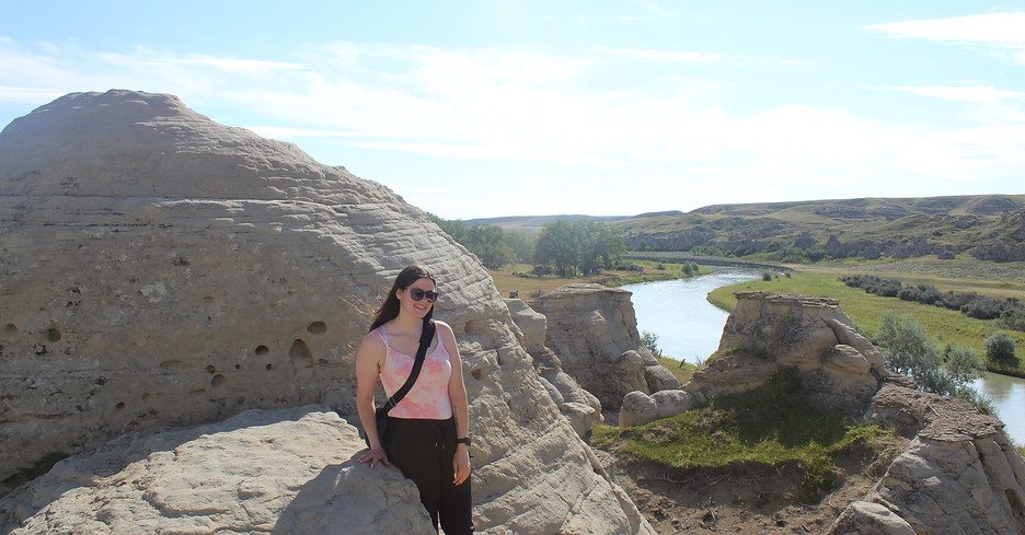 A young women smiling, leaning on a boulder with a grassy scenic background
