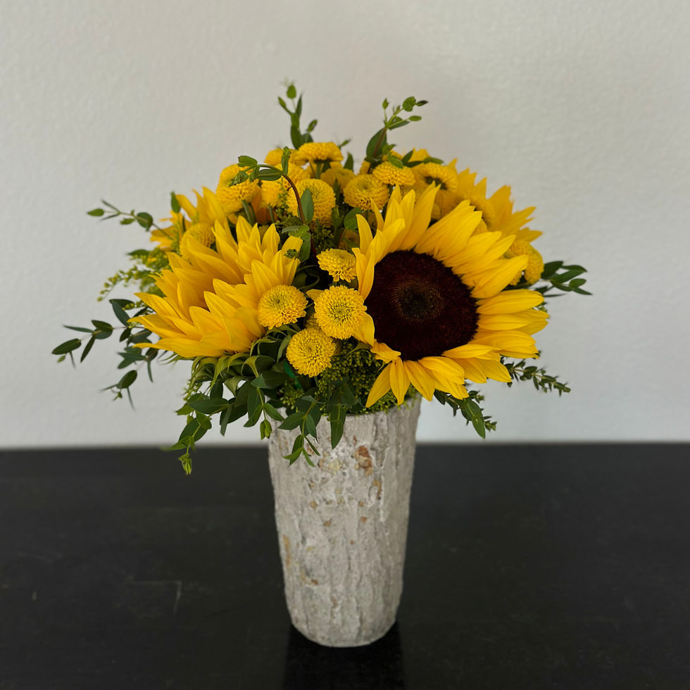 Rustic stone vase filled with bright yellow sunflowers and button poms, arranged with fresh greenery