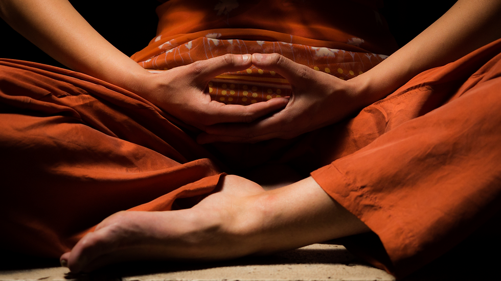 Person in orange robes sits cross-legged in meditation, hands forming a mudra against a dark background.