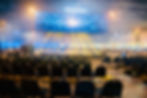 Large audience attending a live corporate event with stage lighting and presentation screen.