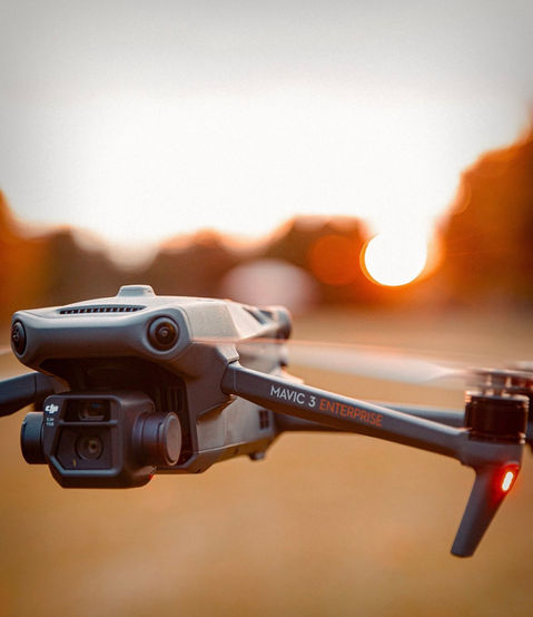 L’image montre un drone DJI Mavic 3 Enterprise en gros plan, avec une mise au point sur son module caméra sophistiqué. Ce module intègre plusieurs capteurs et objectifs, entourés d’un design angulaire. L’un des bras du drone est visible sur le côté, portant l’inscription “MAVIC 3 enterprise”. L’arrière-plan est flou, avec des teintes chaudes et orangées, mettant en valeur les détails du drone.