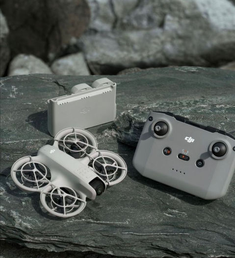 Drone DJI Neo blanc avec protections d’hélices, une radiocommande DJI grise et un hub de chargement pour les batteries, posés sur une roche en extérieur, avec un arrière-plan naturel flou.