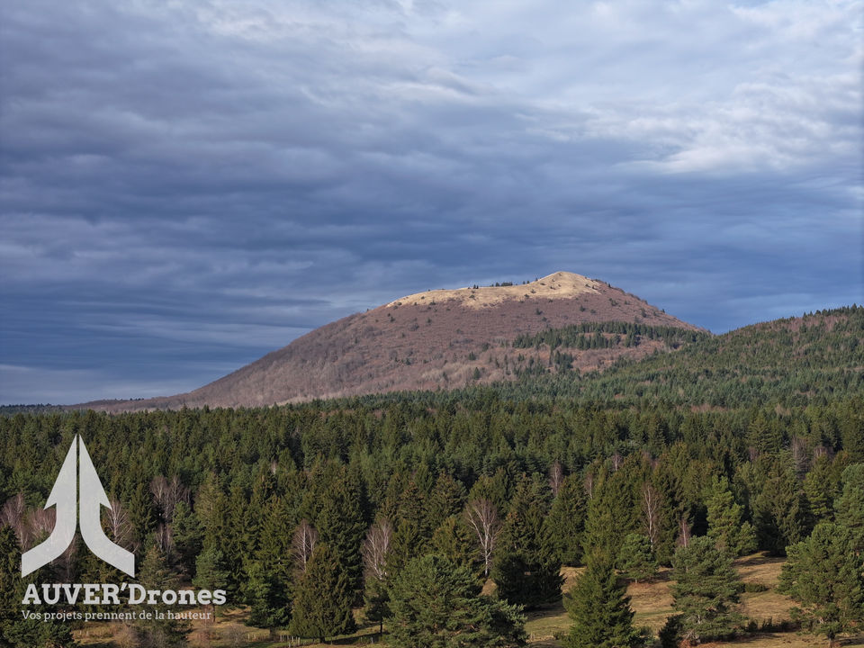 Vue aérienne d’un paysage naturel capturé par Auver’drones, montrant le puy de Come recouvert de végétation dense sous un ciel nuageux. Au premier plan, une forêt de pins s’étend vers la base du volcan. Le logo d’Auver’drones et le slogan « Vos projets prennent de la hauteur » apparaissent en bas à gauche de l’image