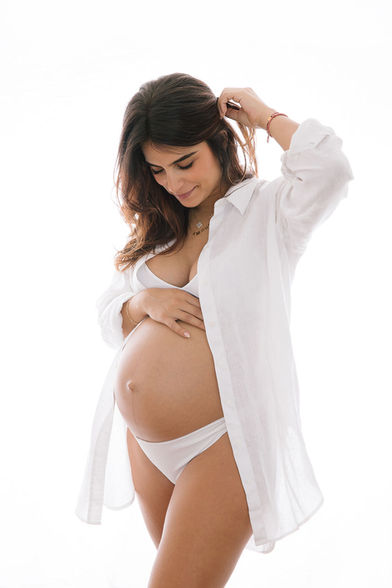 Portrait lumineux et intime réalisé en studio photo à domicile, mettant en vedette une femme enceinte caressant doucement son ventre, incarnant la beauté sereine de la maternité.