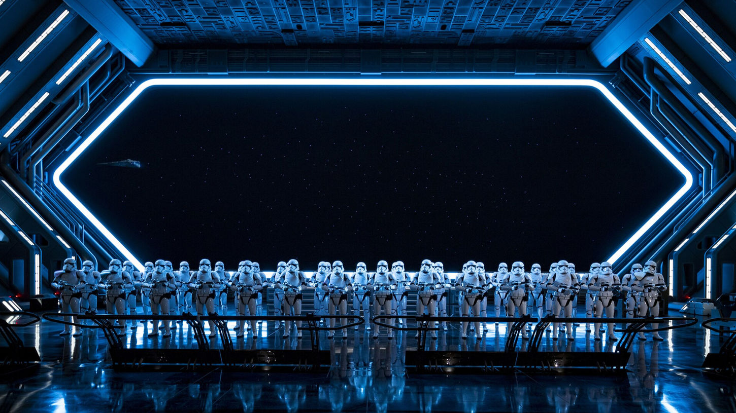 The hangar bay immersive experience from the themed attraction, Star Wars: Rise of the Resistance at Disney World and Disneyland, consisting of metal-clad bent walls with integrated lighting, a large screen behind, a ceiling with a relief of buttons and other objects, and rows of Storm Troopers on the ground behind a barrier rail.
