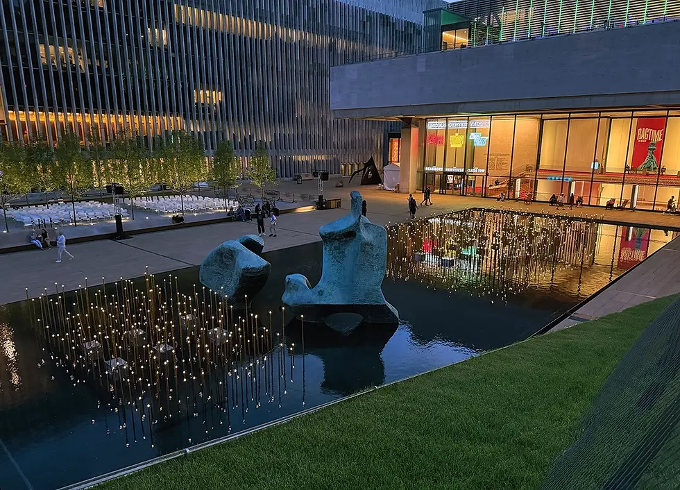 Lincoln Center's Paul Milstein Pool and Terrace at dusk, with a temporary art and light installation of golden rods with lightbulbs on top, and the Lincoln Center Theater behind. 