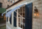 Exterior detail of the Tommy Hilfiger Prep World Cape Cod-style cottage pop-up retail activation, showing the shingles with a scenic weathered finish, windows with navy blue shutters, and custom white and blue stripe fabric awnings.
