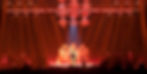 Florence Welch of Florence + The Machine performs at the center of a stage, beneath a large header frame and chandeliers draped in fabric and lights, and in front of a platform with candelabras on stands and draped in fabric and lights, part of the set of Florence + The Machine's North American Dance Fever Tour.