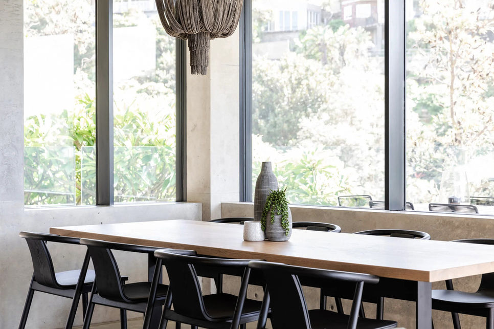 Dining room with concrete walls, timber table, black chairs, woven pendant lighting, and full-height glazing to coastal garden.
