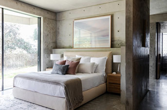 Minimal bedroom with concrete walls, built-in timber shelves, layered bedding, and large window filtered by soft curtains.