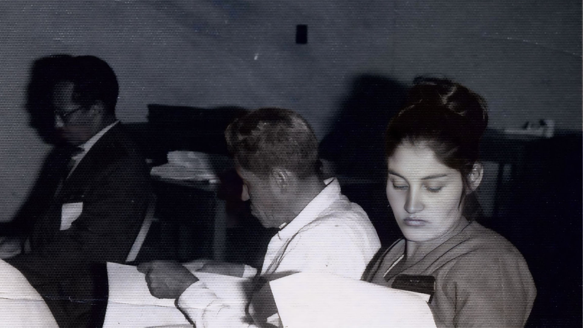 Black and white photo of Doña Angelita sitting in a classroom during a formal meeting, reading documents. This moment is part of her story in Part One of Invisible Grounds.