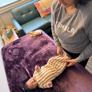 parent in a grey top and dark hair dressing a baby on a purple blanket in a striped gown