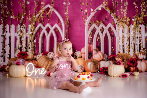 Toddler tasting her cake against a pink and glittery, pumpkin filled backdrop for her First Birthday portraits by Milk & Honey Photo Co in Northwest Indiana.
