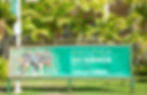 Photo features a large, bright, grass green banner hanging in front of a campus building, the view of which is obscured by large palm fronds. On the banner, four traditionally-aged, diverse students are sitting next to each other smiling. Two girls, both with sunglasses perched atop their heads, sit in between two boys, both of whom are using their right hands to gesture the Hawaiian "shaka" sign. Text on the banner next to the students reads: "Discover UH Manoa Open House This Saturday."