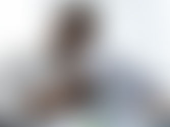 An African-American man wearing a white dress shirt and glasses is reading a piece of paper with a green sticky note in his hand while sitting at a desk with a laptop. Large windows are behind him creating a bright and well-lit work atmosphere.