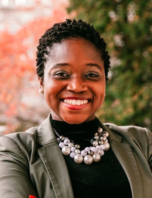 Smiling person with short hair in a blazer and beaded necklace stands outdoors. Autumn leaves and greenery visible in the background.