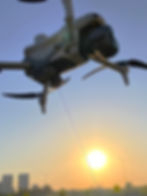 Drone flying at sunset against a clear sky. A thin wire is visible from the drone. Silhouetted city skyline below, warm sunlight.