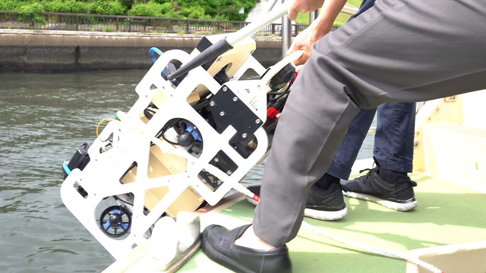 Two people on a boat lower a white robotic device into the water. One person wears black shoes. The background shows calm water and greenery.