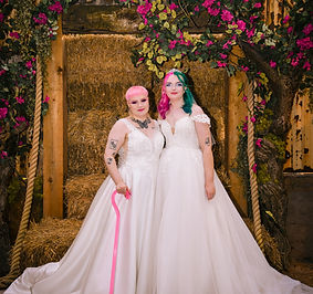 Two people stand side by side in matching white wedding gowns, posing beneath an archway covered in lush greenery and bright pink flowers. One has short pink hair and holds a pink cane, while the other has long hair in shades of teal and magenta, wearing a tiara. Behind them are stacked hay bales and rustic wooden beams, creating a warm, whimsical backdrop. Both look directly at the camera with calm, happy expressions.