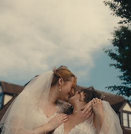 Two grooms in suits are laughing and holding hands outside of a brick building. There are wedding guests around them, throwing confetti.