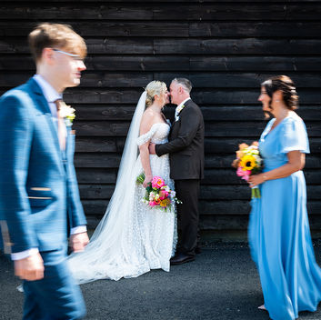 Wedding couple kissing in front of a black wooden wall, with blurred guests in blue passing by. Photo is by Kiss Photo - a LGBTQ+ Inclusive Wedding Photographer