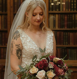 A to-be-wed wearing a beaded lace wedding dress and a long veil stands in front of tall wooden bookshelves filled with books. She looks down at a bouquet of flowers with greenery, and a tattoo is visible on her upper arm.