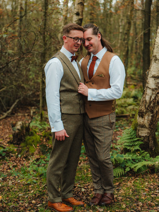 A newly wed couple stand in a green mossy forest on their wedding day. They intimately embrace. The person on the right has a long, low ponytail and is wearing a light brown waistcoat. Whilst the person on the left has short brown hair and wears glasses and is wearing a light green waistcoat. Their heads press together. Image By: Eli Wing Photography
