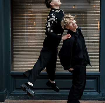 A non-traditional wedding scene with one person in a wedding dress and makeup sitting, while another adjusts their veil, against a backdrop of a whimsical painting