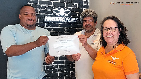 Três pessoas, sendo um aluno e dois instrutores da Escola de Drone da Bahia, sorriem enquanto seguram um certificado de conclusão do curso de piloto de drone presencial em Salvador.