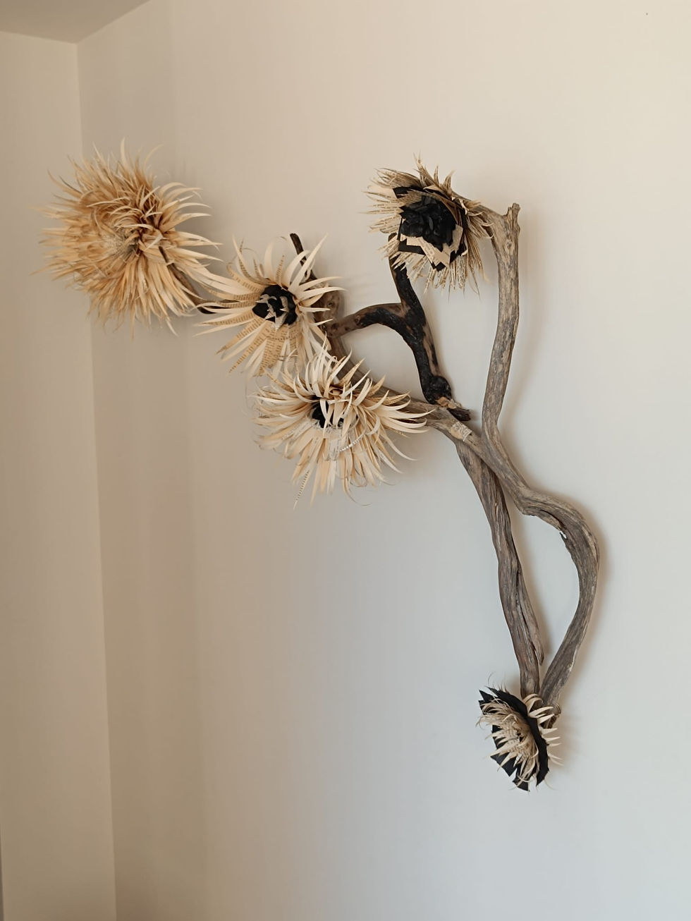 Sculpture murale nommée Lisea, composée de morceaux de bois flotté grisés et noircis par le feu, ornée de fleurs en papier de livres recyclés, fixée sur un mur clair.