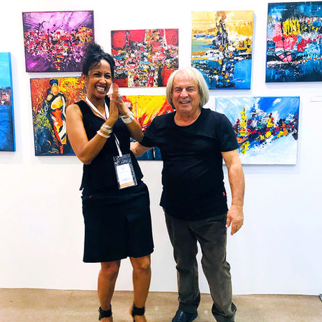 Artist Arlia-Hamda Elmi sharing a joyful moment on the exhibition floor next to her colorful abstract artworks with a fellow artist and friend.