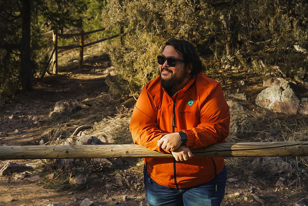 Photographer Matt Archuleta wearing an orange jacket leaning on a wooden railing along a wooded trail in natural light.
