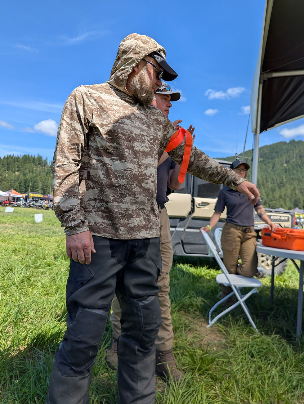 Man in camo hoodie and cap gestures beside others at an outdoor grassy event with mountains, blue sky, and a vehicle nearby.