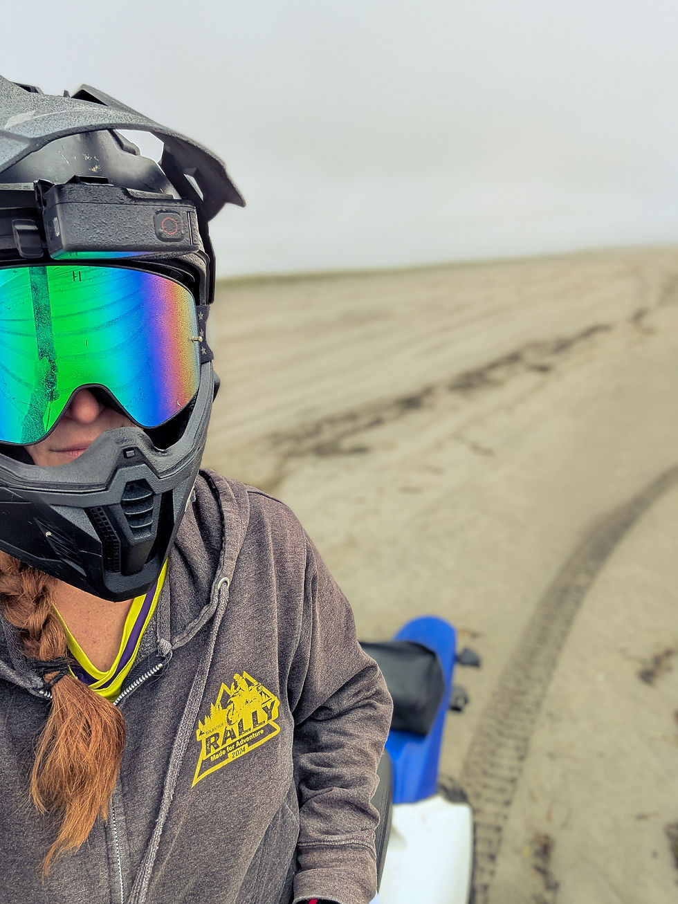 Helmeted ATV rider with rainbow visor in a sandy beach, gray hoodie