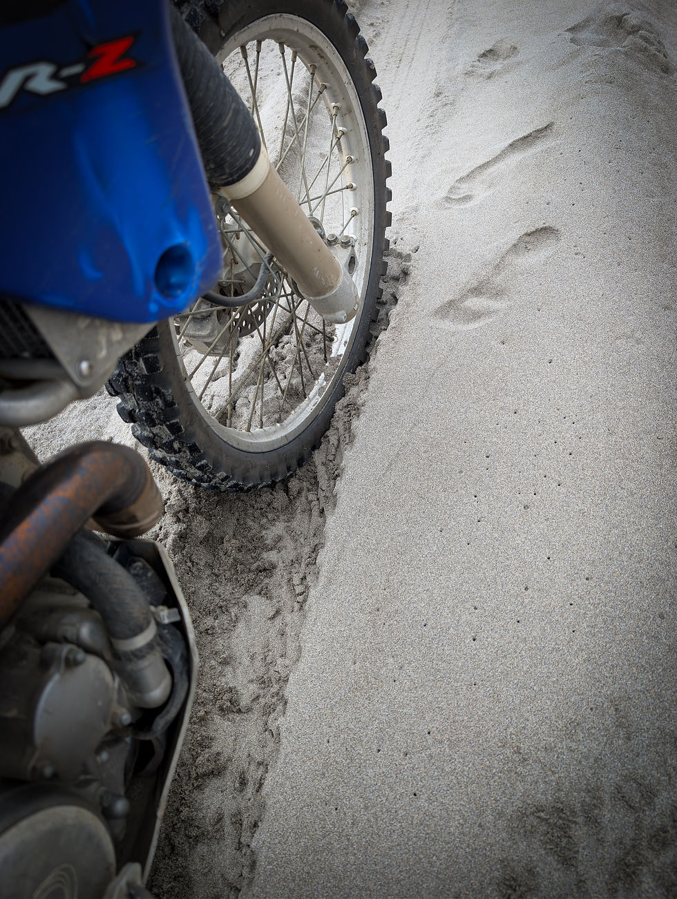 Close-up of a blue dirt bike wheel sunk in soft sand, with tire tracks and the text DRZ on the fairing.