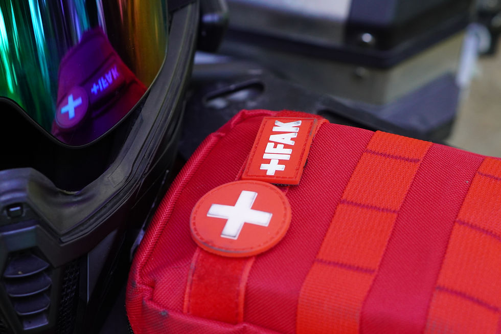 Close-up of a black helmet beside a red medical pouch with white cross and HFAK patch, with rainbow visor reflection.