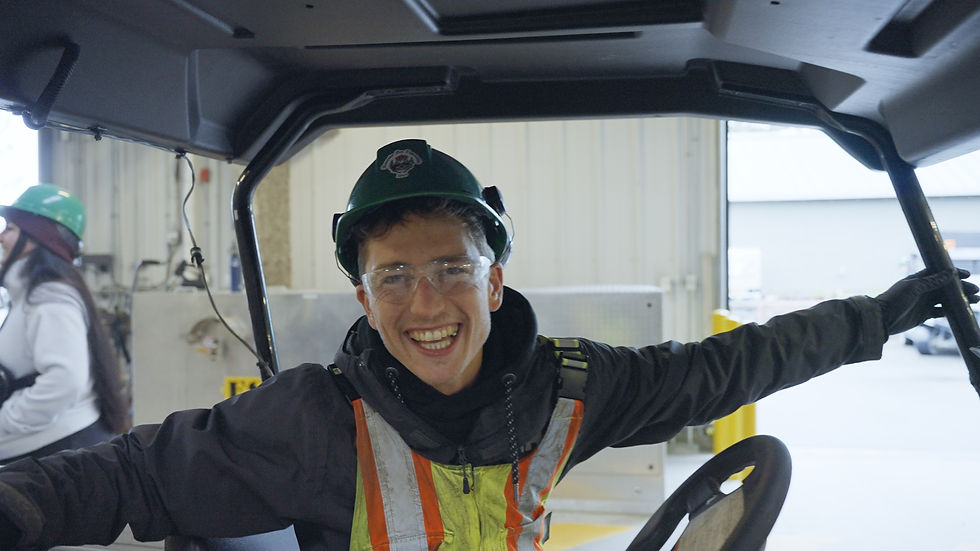 Person smiling inside a golf cart in an industrial setting, wearing a green hard hat and safety gear.
