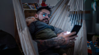 A child sleeping on a parents shoulder in the dark, while they look at a smart phone.