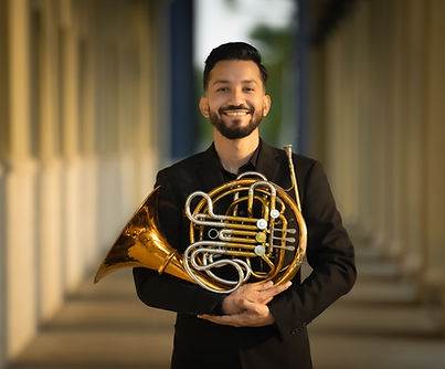 Alex Sanchez in a black suit, holding a French horn, while smiling at the camera. 