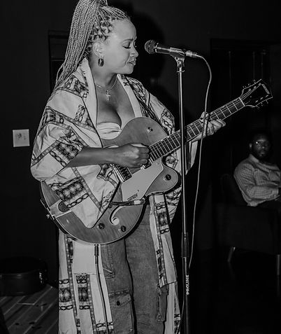 A woman playing an electric guitar in a dimly lit room, wearing a patterned coat and braids. She is singing into a microphone. In the background, a person is seated, watching her performance.