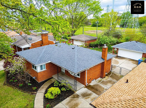 Aerial view of a house with a new gray shingle roof, red brick exterior, and two brick chimneys—showcasing expert roofing services—surrounded by trees, a driveway, and nearby garages.